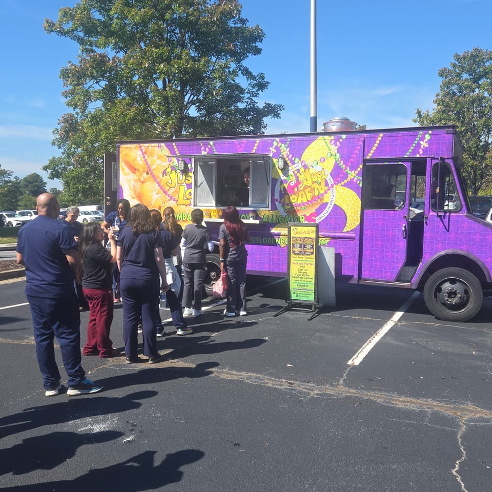 Customers queue at a brightly decorated purple seafood food truck in a sunny parking lot.